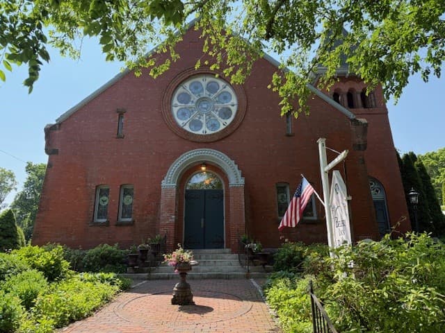 A red brick building with a large round window, surrounded by greenery and featuring a sign and American flag.