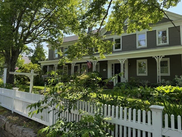 A charming two-story house with a white picket fence and lush greenery, featuring an American flag.