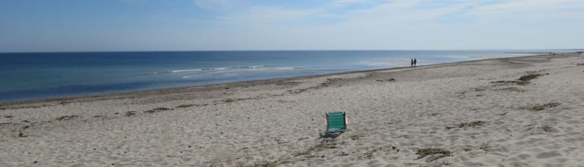 A solitary beach chair on a sandy shore with distant figures walking along the water's edge.
