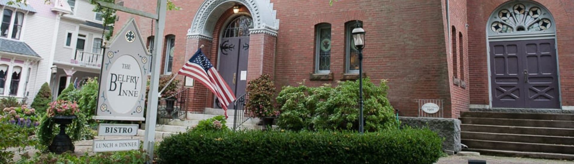The entrance of The Belfry Inn featuring a sign, landscaped greenery, and an American flag.