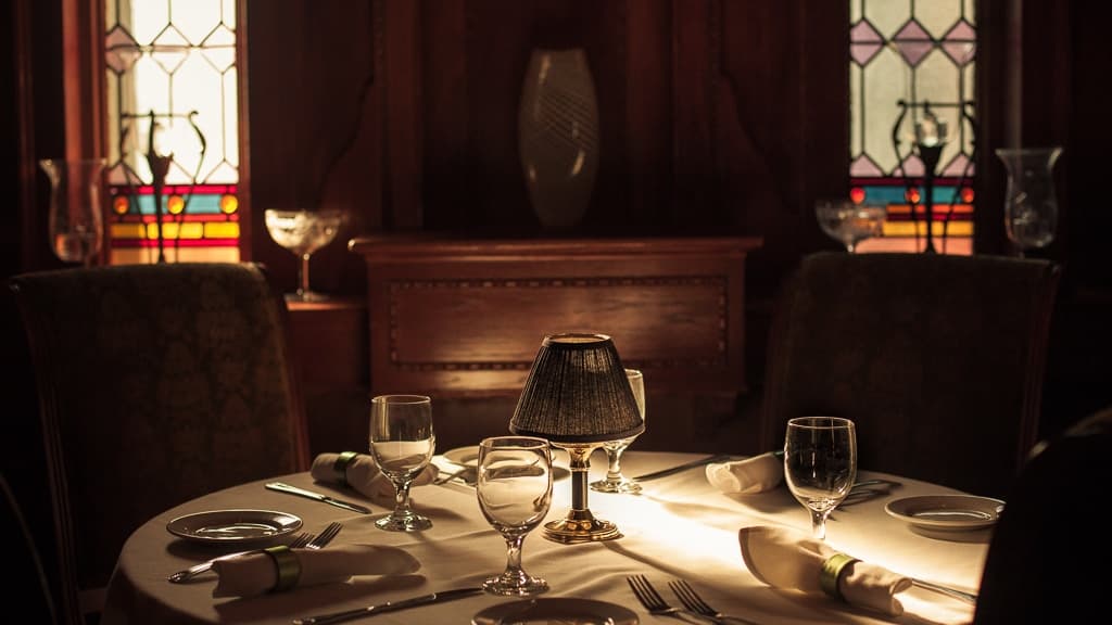 A beautifully set dining table with elegant glassware and muted lighting, framed by stained glass windows.