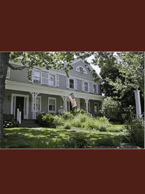 Exterior of a federal style home from 1827 with grey siding and a US Flag surrounded by trees