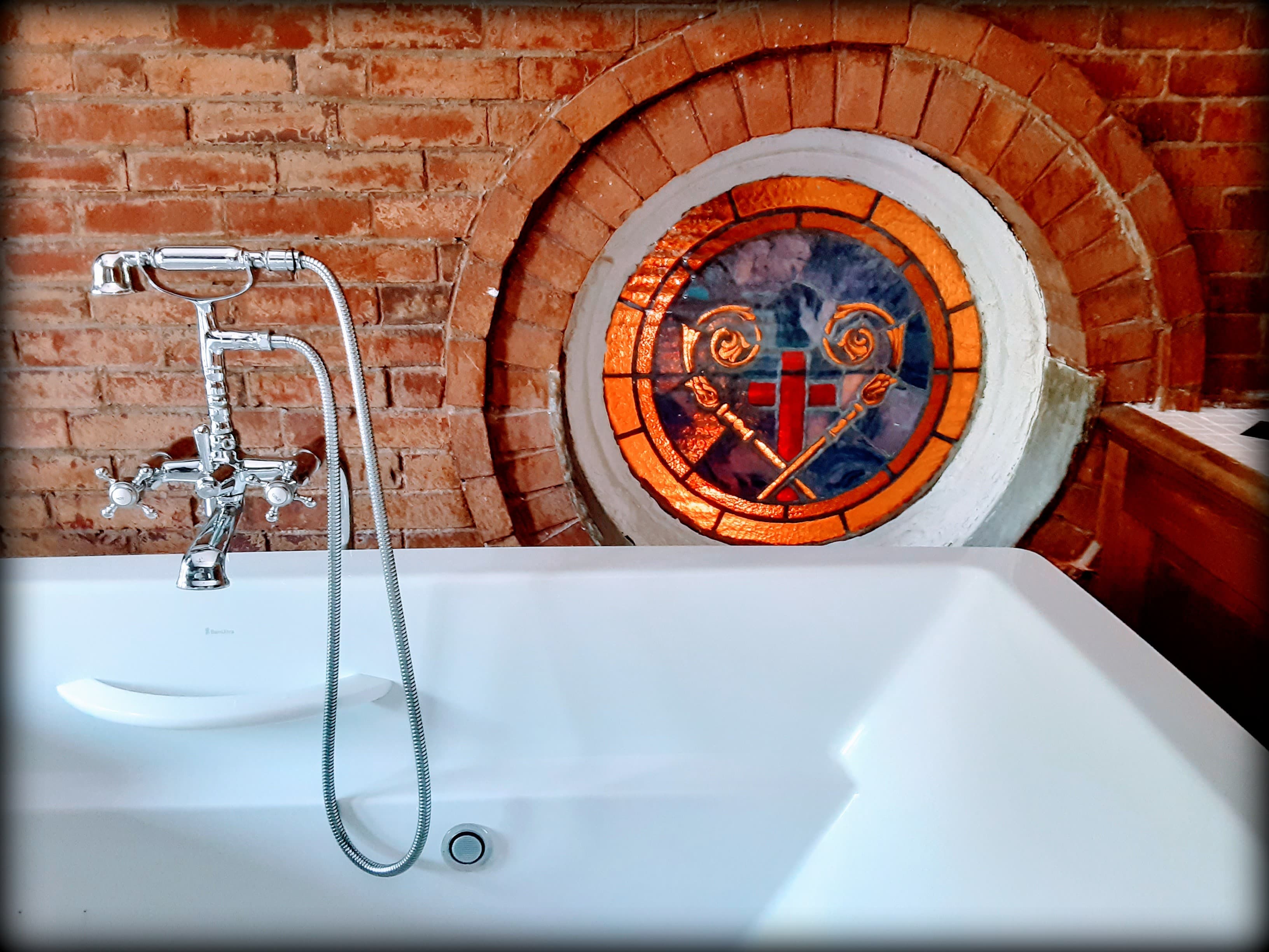 A modern white bathtub in front of a circular stained glass window set in a brick wall.