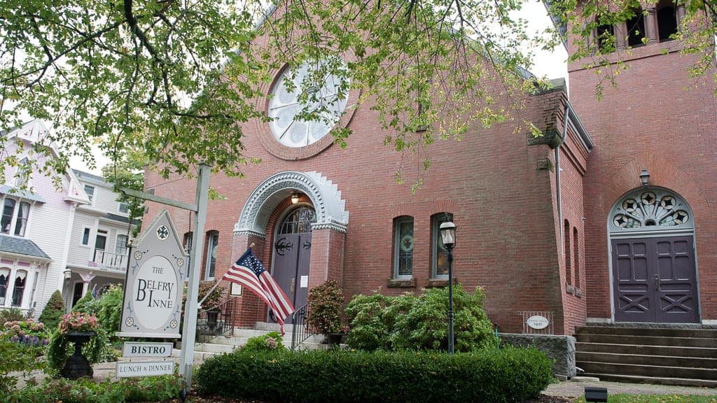 A brick building with a distinctive circular window and a sign for "The Belfry Inn Bistro" surrounded by greenery.