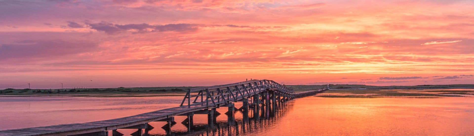 A wooden bridge stretches over a calm waterway under a vibrant pink and orange sunset sky.