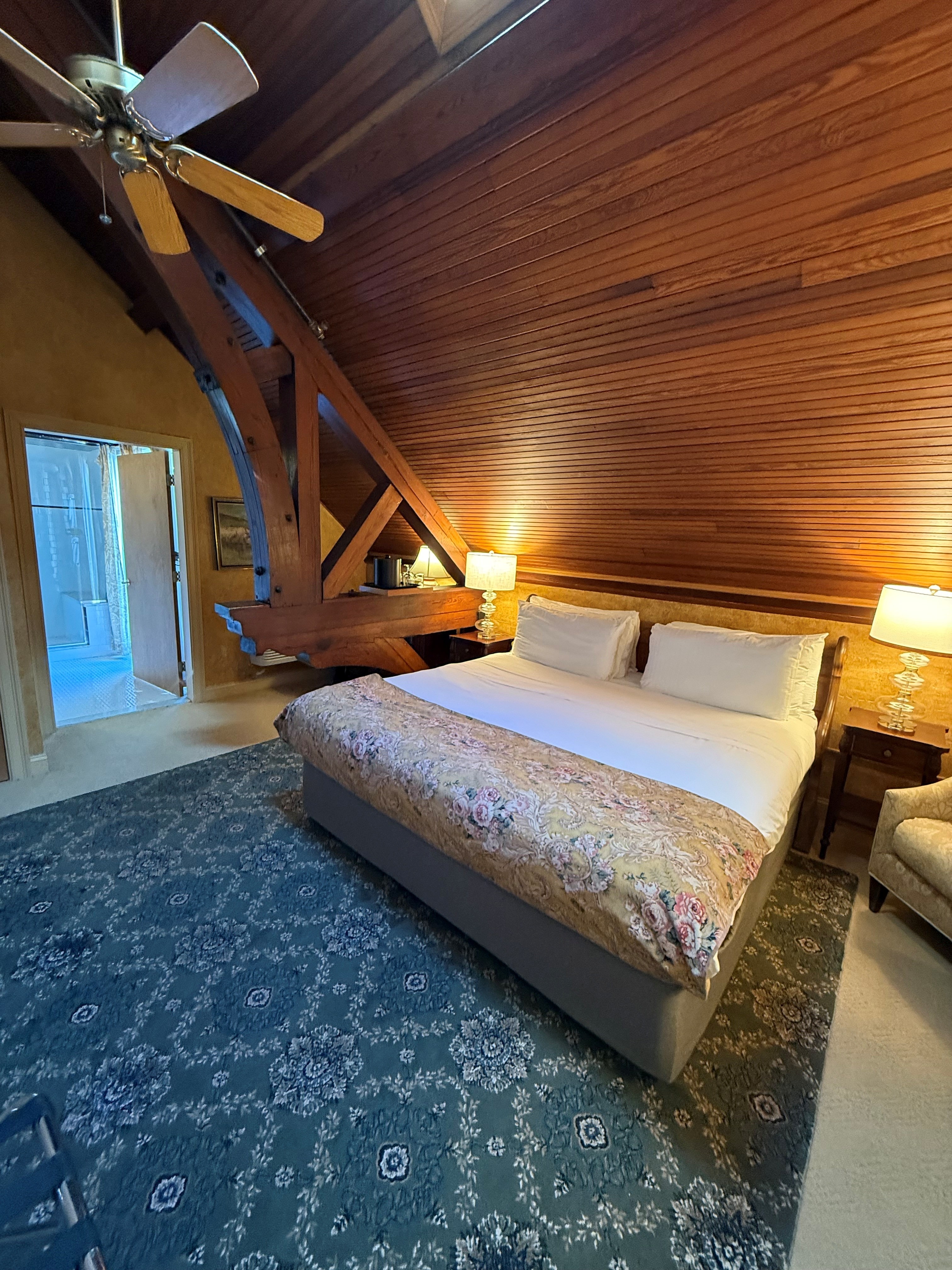 A cozy bedroom with a sloped wooden ceiling, featuring a large bed, decorative rug, and two lamps.