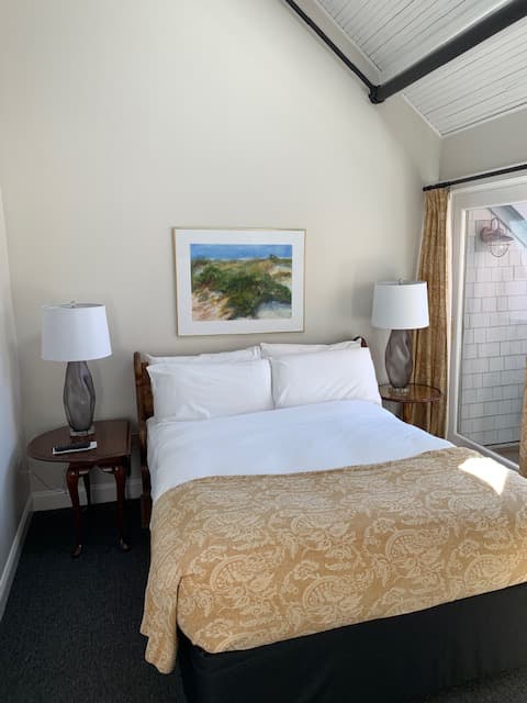 A cozy bedroom featuring a bed with white linens and a decorative gold blanket, flanked by two lamps and adorned with a landscape painting on the wall.